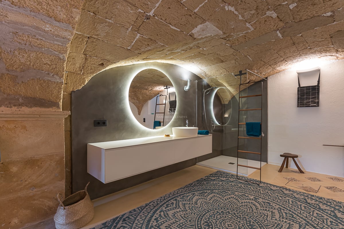 Salle de bain avec plafond voûté en pierre, miroir rétroéclairé et meuble vasque suspendu blanc.