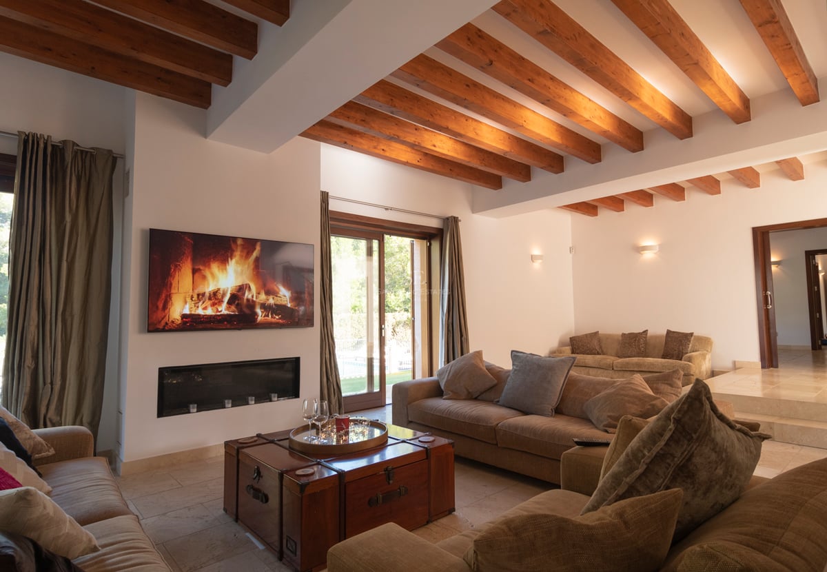 Vardagsrum med takbjälkar i trä, modern öppen spis, stengolv och stora glasdörrar mot trädgården.