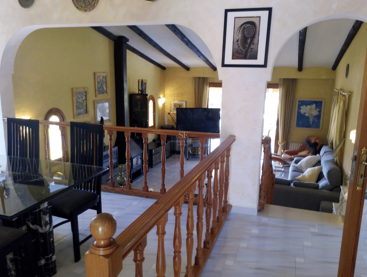 Salón a dos niveles con suelos de mármol, barandilla de madera y vigas oscuras bajo arcos blancos.