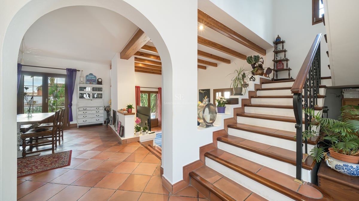 Interior view showing terracotta floors, an arched entrance, wooden ceiling beams, and a staircase with iron railings.