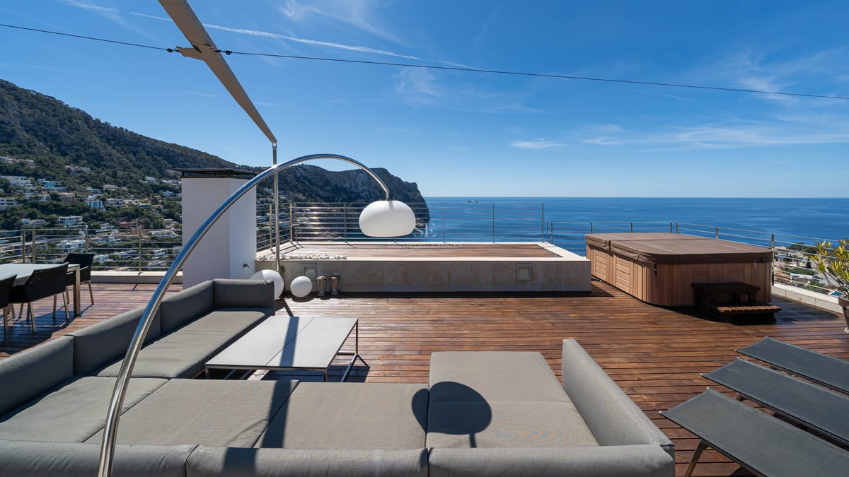 Modern rooftop terrace with grey outdoor sofa, wooden decking, and a hot tub overlooking the blue sea and mountains.