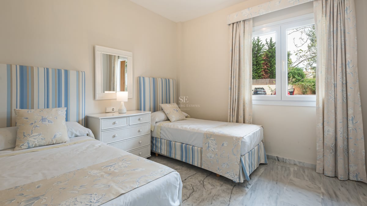 Sunlight-filled twin bedroom featuring blue and white linens, white dresser, and polished marble floors.