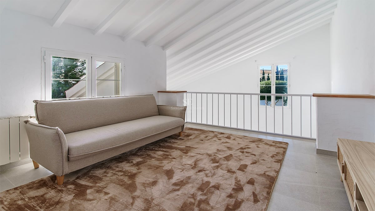 Vardagsrum på loftet med beige soffa, mönstrad brun matta och vitmålade takbjälkar i trä.