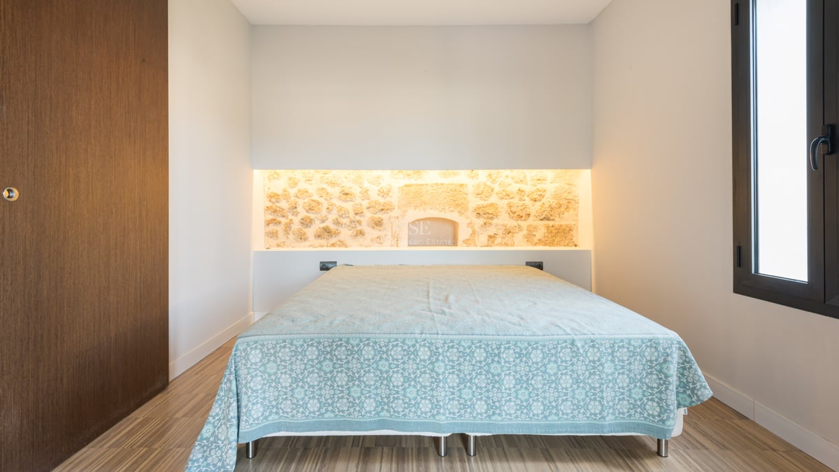 Sovrum med en säng, en belyst stenvägg i bakgrunden och ljust trägolv.
