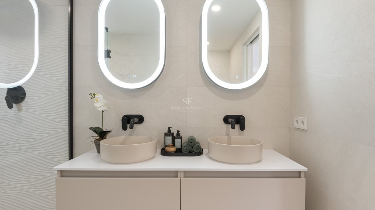 Modern bathroom with two round vessel sinks, black faucets, and oval backlit mirrors on a beige wall.