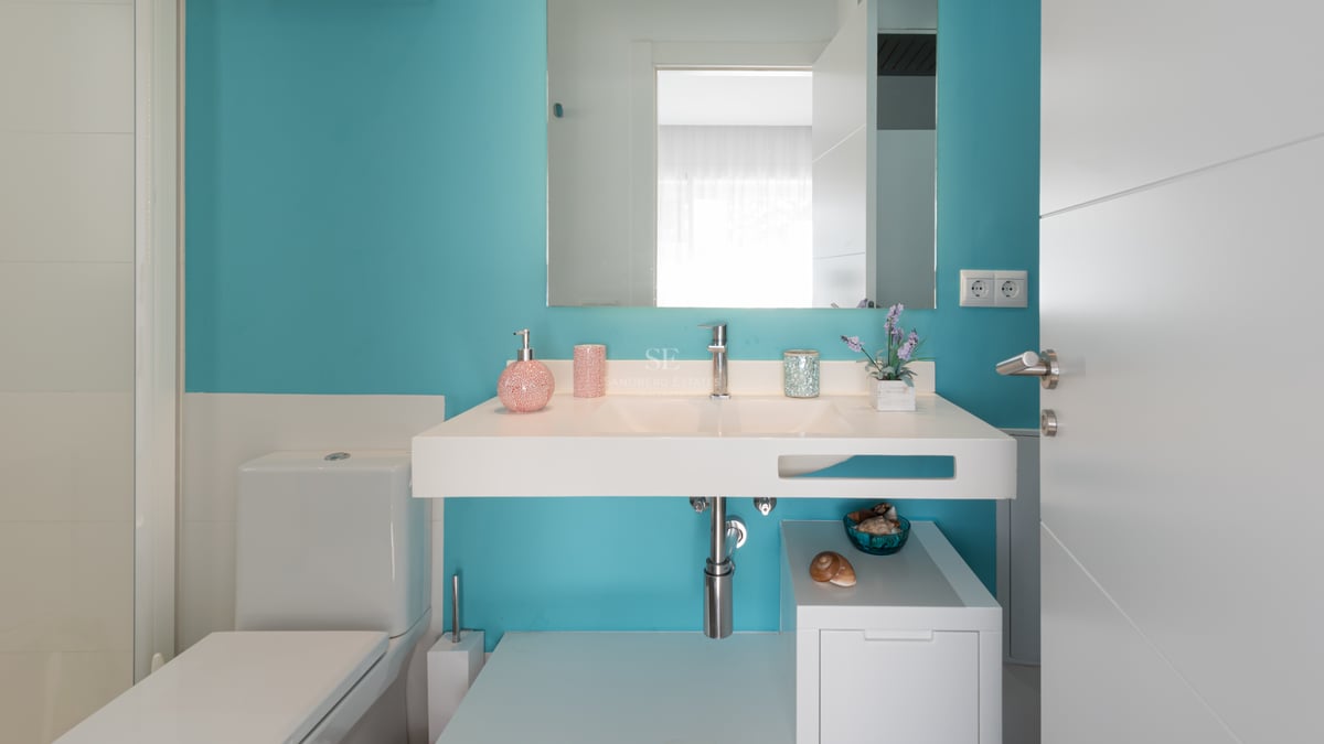 Salle de bain moderne avec mur turquoise, vasque suspendue blanche, grand miroir et toilettes blanches.