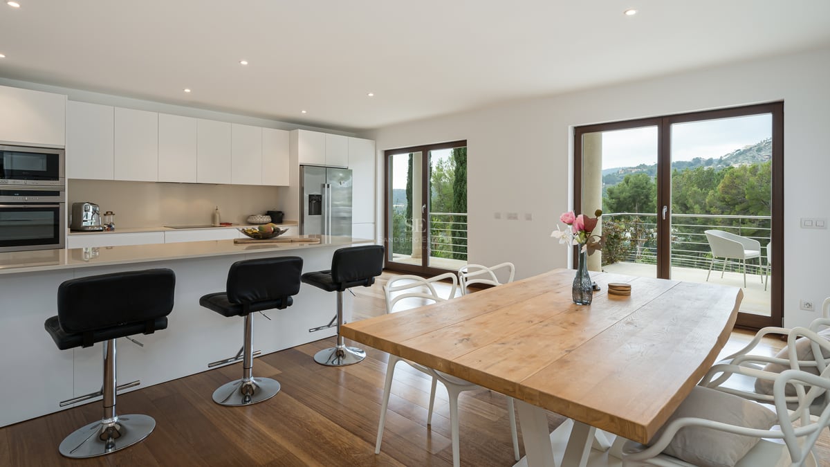 Cuisine contemporaine blanche avec îlot central, table de repas en bois et baies vitrées ouvrant sur la nature.