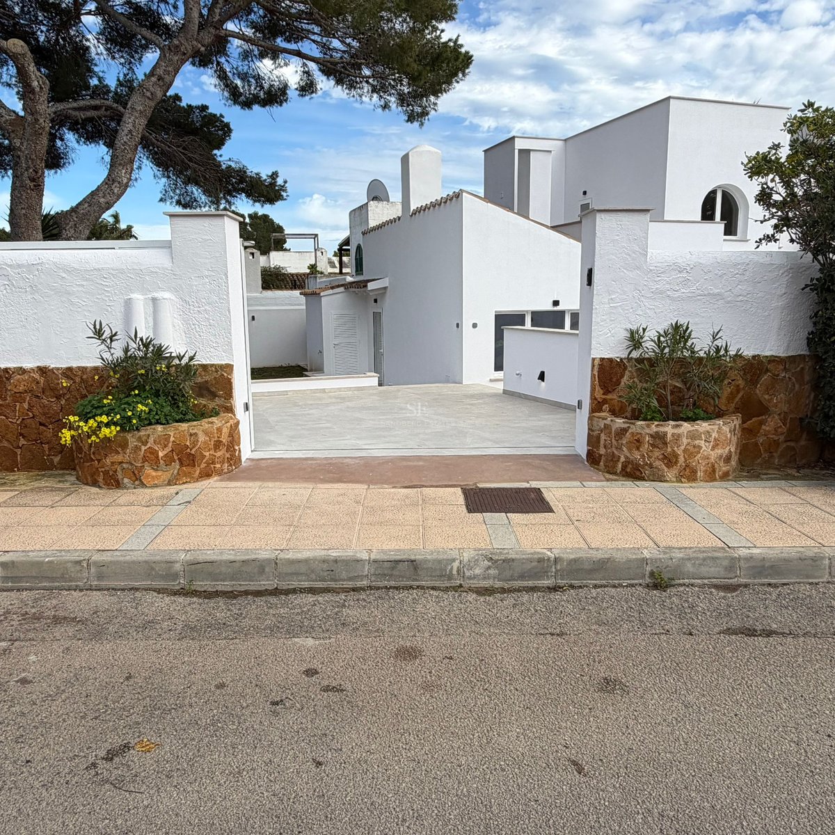 Entrance to a white villa featuring stone-clad walls, planters with yellow flowers, and a paved driveway under pine trees.