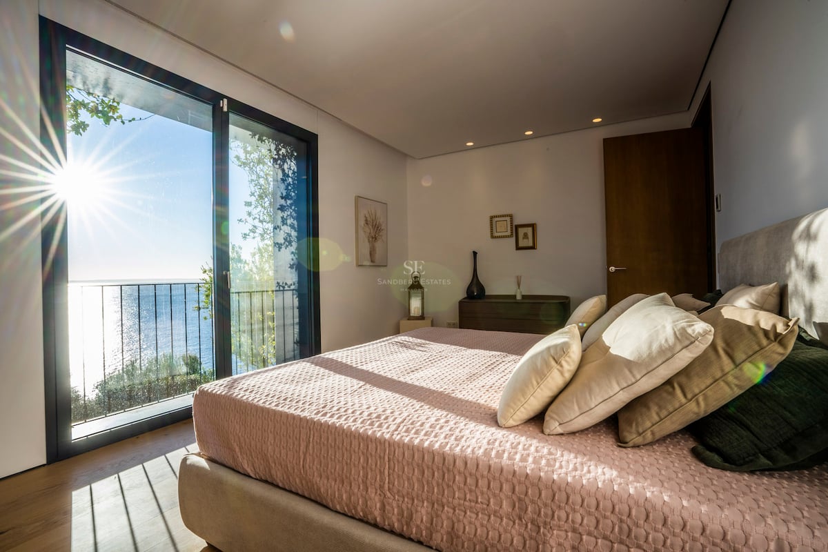 Helles Schlafzimmer mit großem Bett, rosa Bettwäsche, Eichenparkett und bodentiefem Fenster mit Blick auf das blaue Meer.
