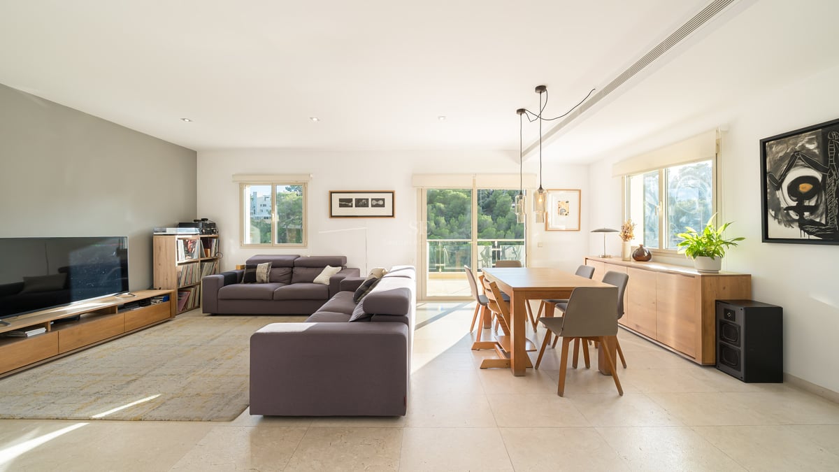 Ljust vardagsrum med grå soffa, matbord i trä och stora fönster som vetter mot en skog.