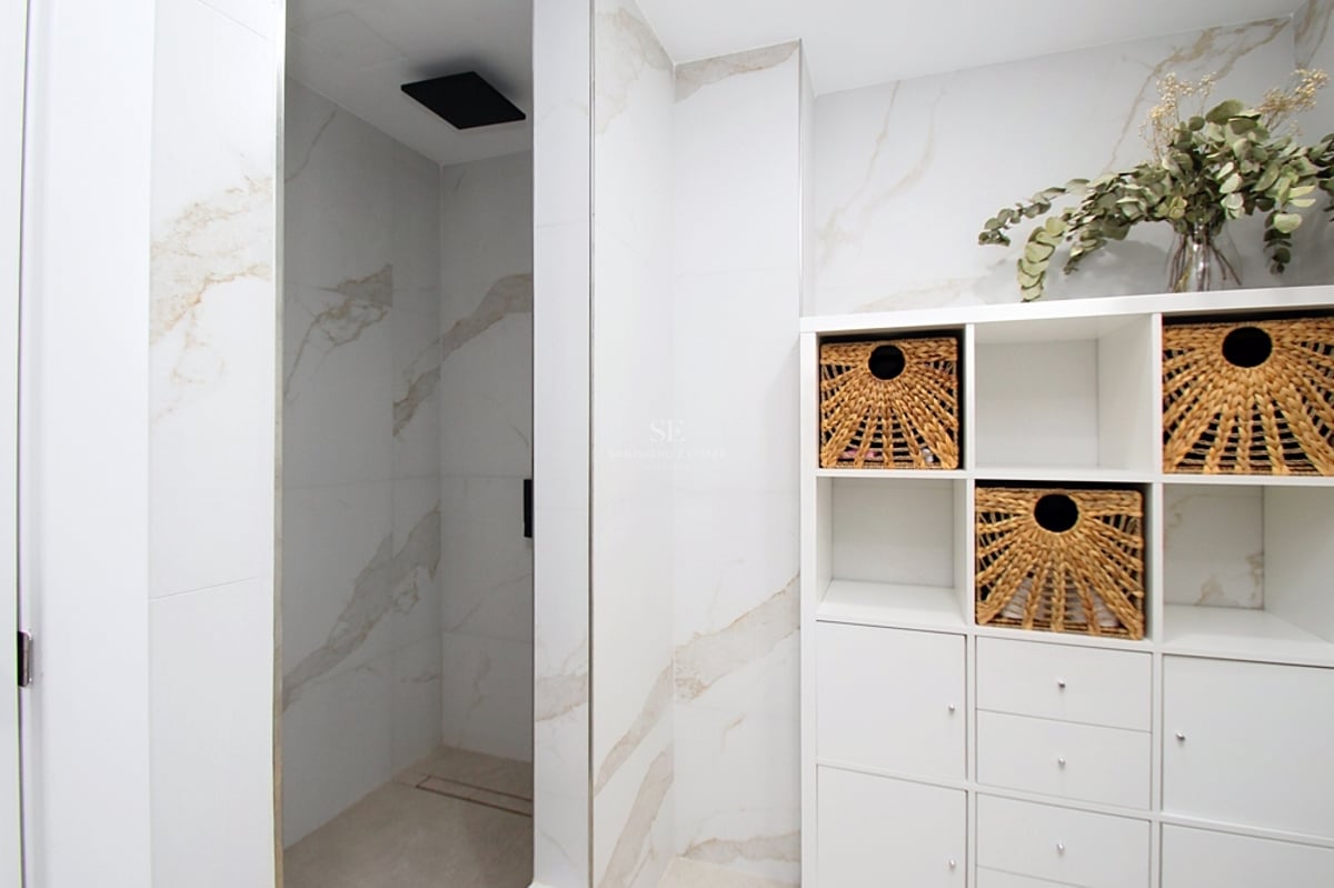 Ett modernt badrum med en rymlig dusch, väggar i marmoreffekt, svart takdusch och vit hylla med korgar.