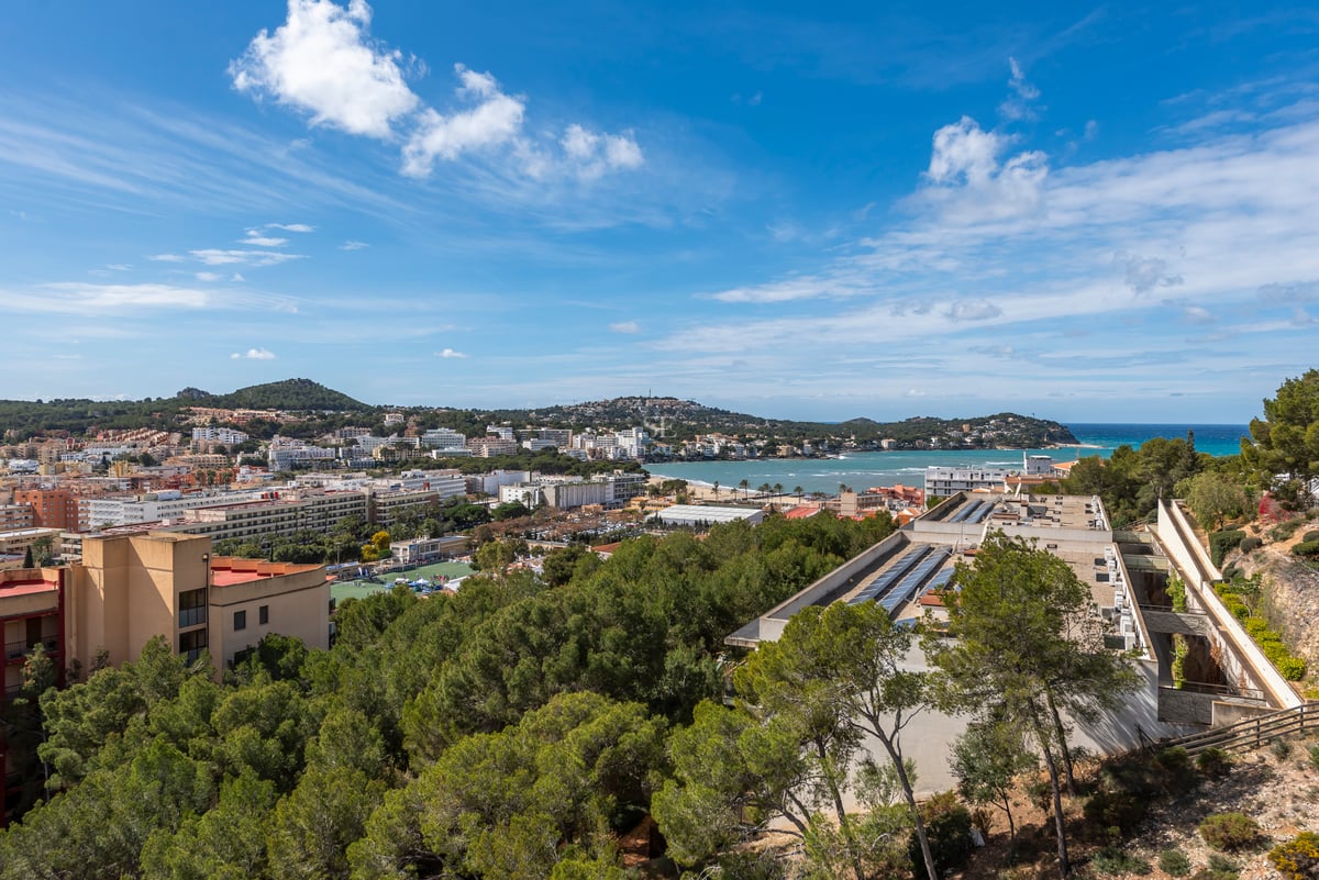 Vue surélevée d'une ville côtière méditerranéenne avec mer bleue, pinèdes et montagnes sous un ciel clair.