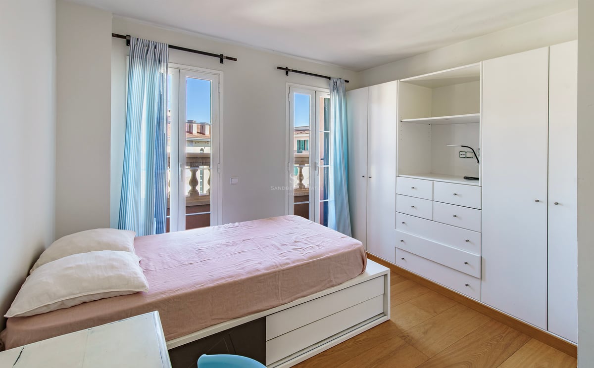 Ljust sovrum med rosa sängkläder, vita garderober och franska dörrar med blårandiga gardiner.