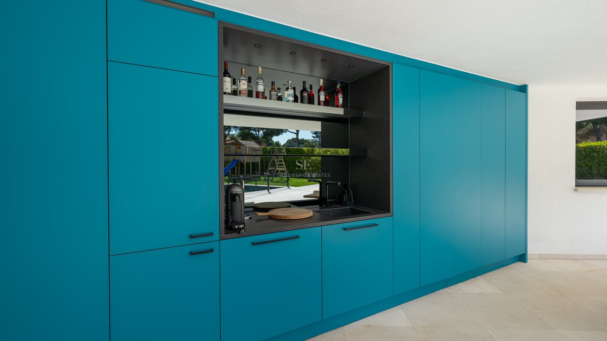 Mobilier de cuisine moderne bleu canard avec bar intégré et reflet du jardin dans la crédence en verre.