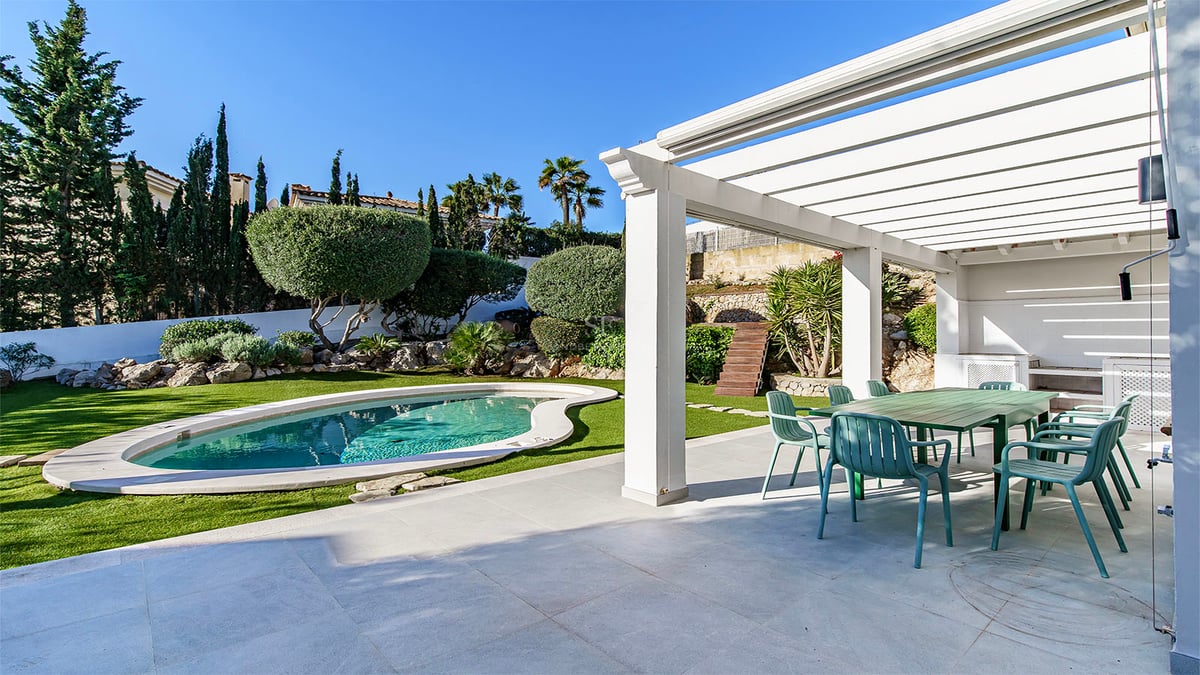 Vit pergola som täcker en matplats utomhus med gröna stolar bredvid en njurformad pool och prunkande trädgård.