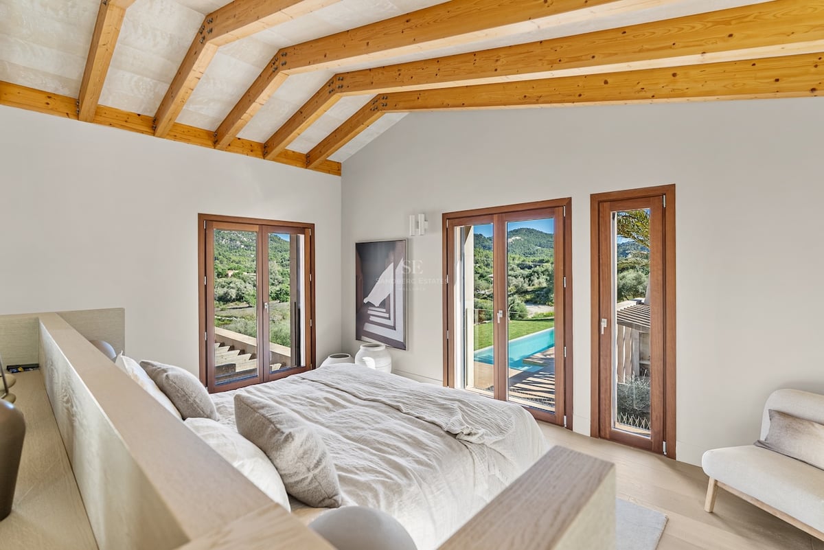 Chambre moderne avec plafond à poutres en bois, lit en lin beige et portes vitrées donnant sur une piscine et collines.