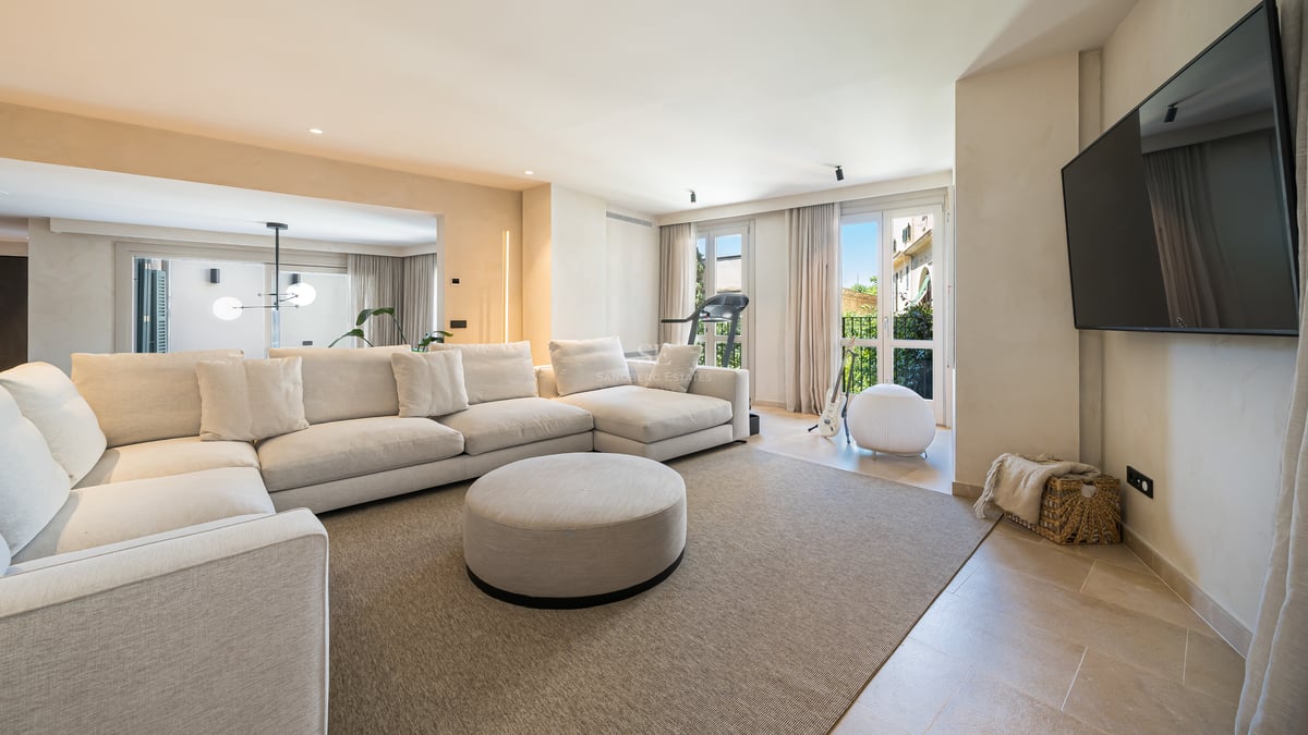 Un salon moderne et lumineux avec un grand canapé d'angle crème, un pouf rond et des sols en pierre naturelle.