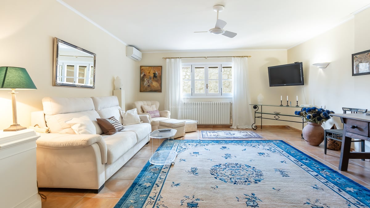 Salon lumineux avec canapé blanc, grand tapis bleu à motifs, sol en terre cuite et ventilateur de plafond.