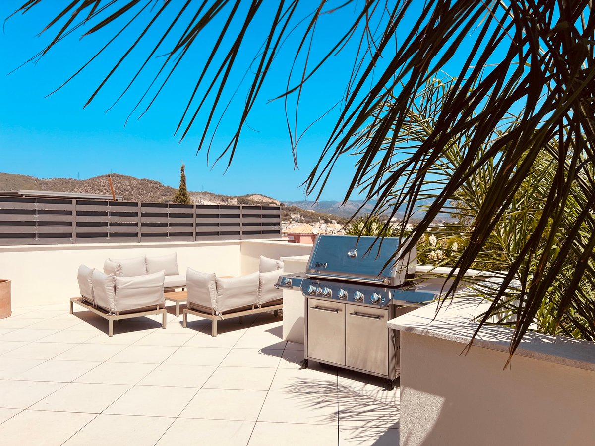 Toit-terrasse ensoleillé avec grand barbecue en inox, salon de jardin moderne et vue sur les montagnes.