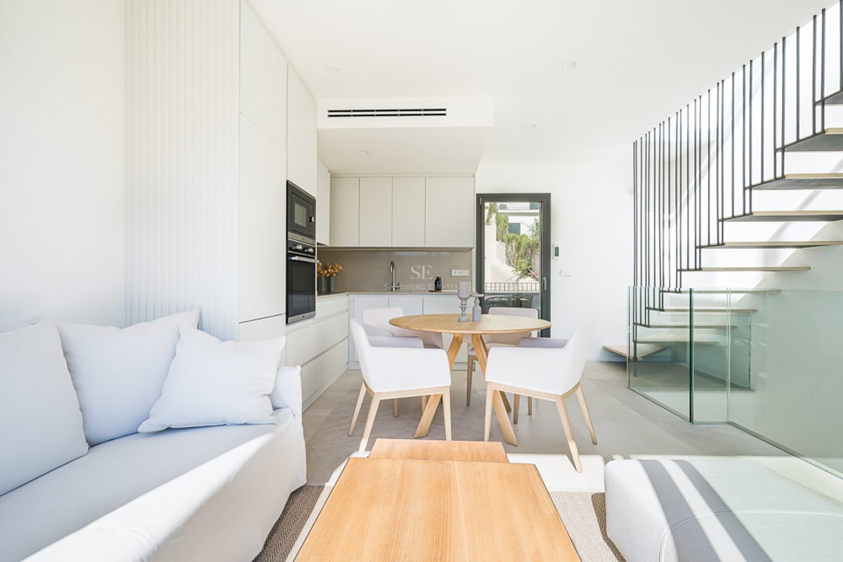 Bright minimalist living room with white sofa, oak dining table, sleek kitchen, and a glass-enclosed floating staircase.
