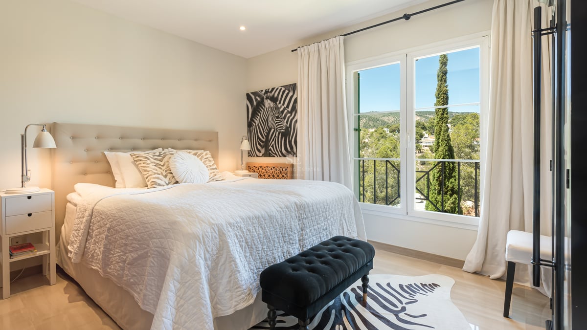 Bright bedroom featuring a large bed with a tufted headboard and a window overlooking lush green hills and a cypress tree.