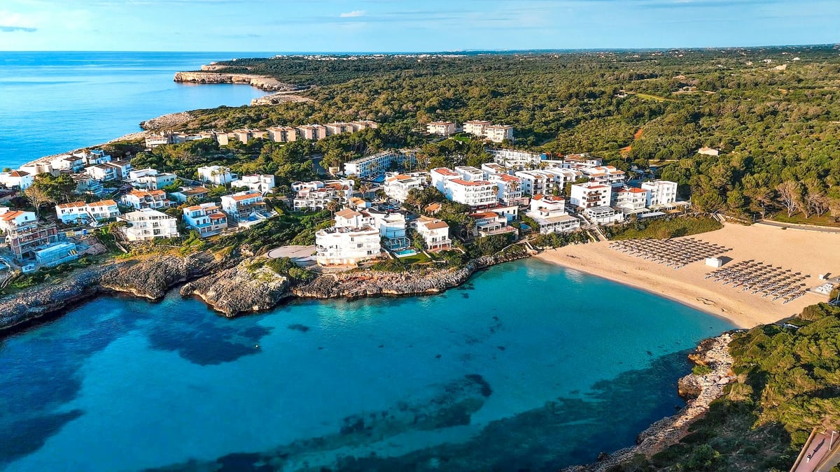 Vue aérienne d'un complexe de bâtiments blancs à côté d'une crique turquoise et d'une plage entourée d'une forêt verte.