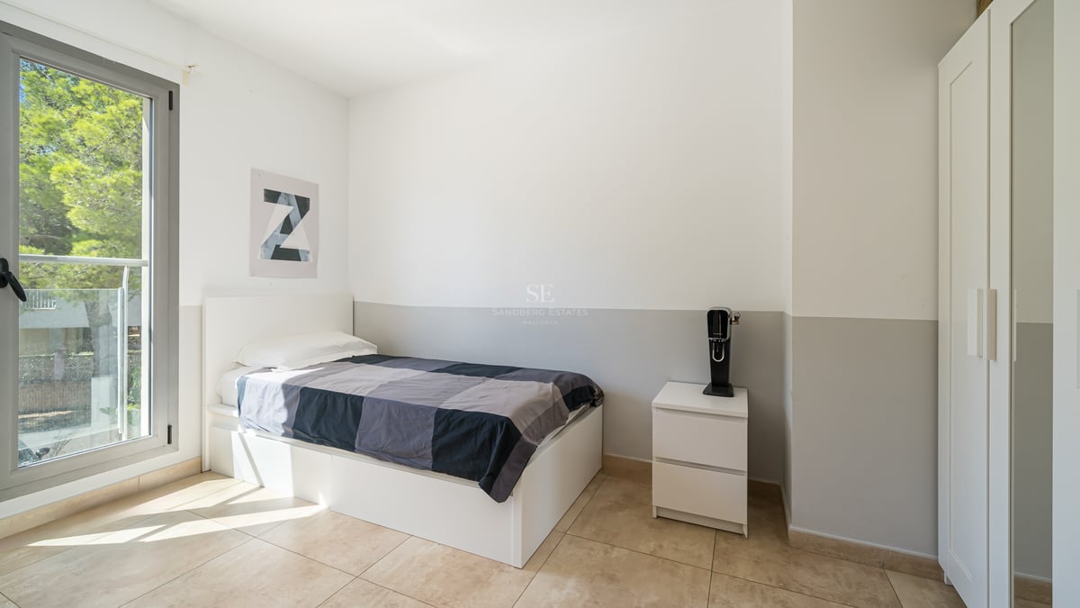 Ett ljust sovrum med en vit enkelsäng, klinkergolv, vit garderob och ett stort fönster med utsikt över träd.