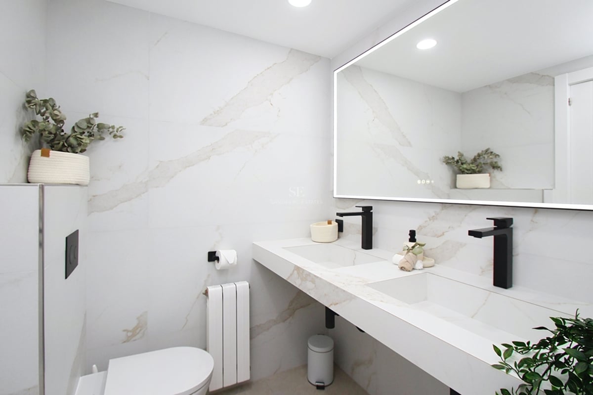 Modernt badrum med vita marmorliknande väggar, dubbeltvättställ med svarta kranar och en stor bakgrundsbelyst spegel.