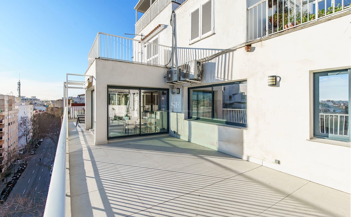 Terrasse ensoleillée avec carrelage clair, garde-corps blancs et vue sur une rue citadine sous un ciel bleu.