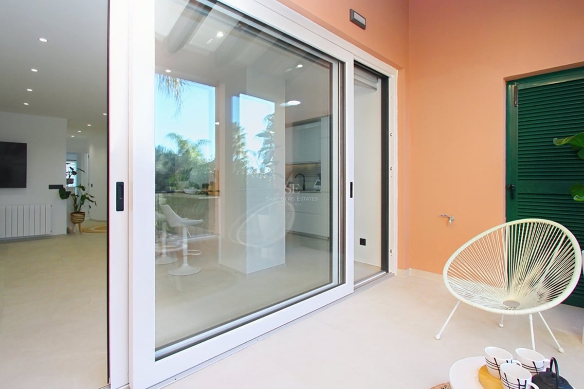 En solig terrass med en vit Acapulco-stol och skjutdörrar i glas som öppnar upp mot ett modernt vardagsrum.