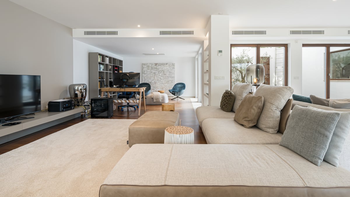 Salon contemporain avec grand canapé en L beige, parquet en bois et coin bureau au fond avec plusieurs écrans.