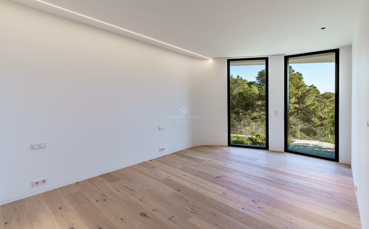Pièce vide lumineuse avec parquet en bois clair, murs blancs et grandes fenêtres donnant sur la forêt.