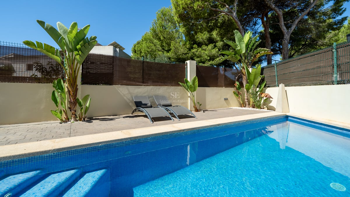 Blå mosaikpool intill en stenlagd terrass med grå solstolar och tropiska växter under en blå himmel.