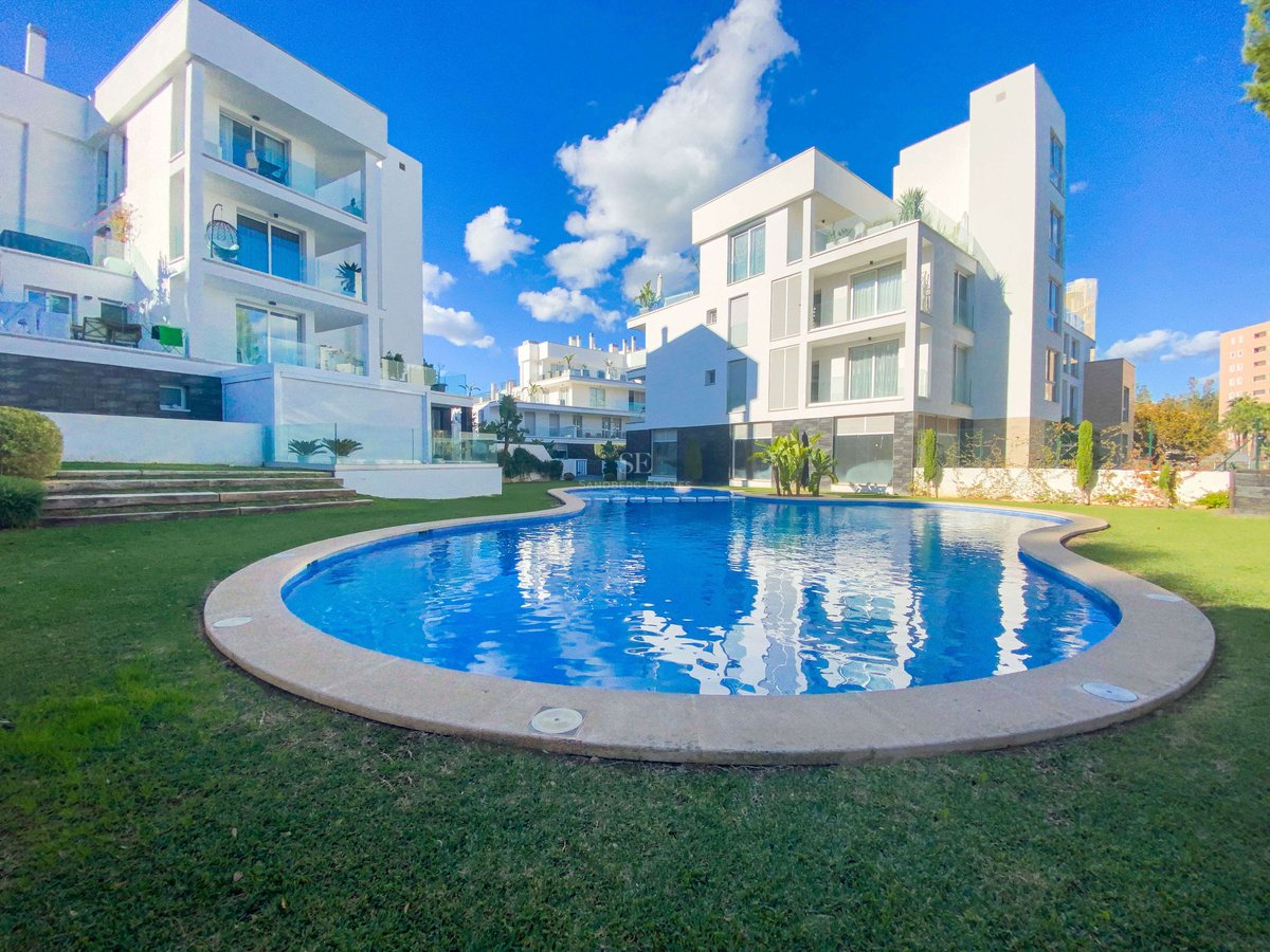 Geschwungener blauer Pool umgeben von Rasen und modernen weißen Gebäuden unter strahlend blauem Himmel.