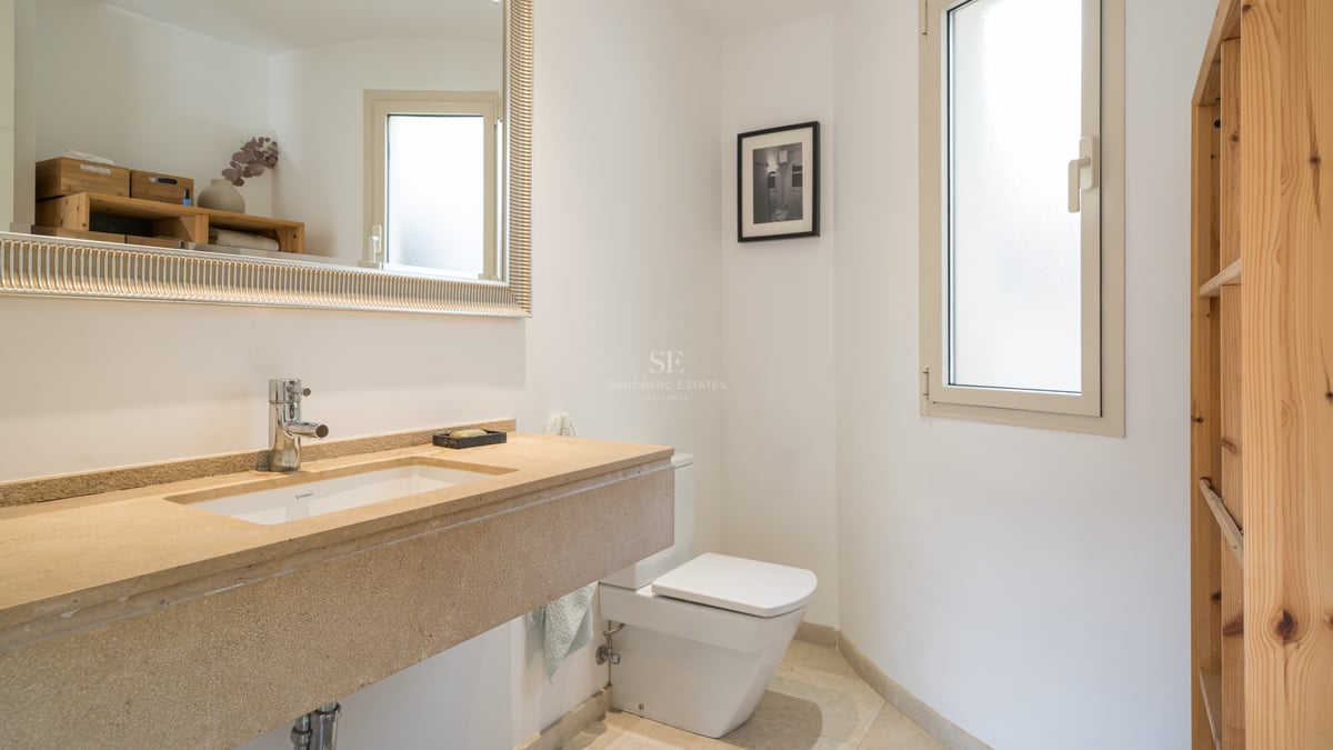 Ljust badrum med tvättställ i beige sten, stor spegel med silverram, vit toalett och frostat fönster.