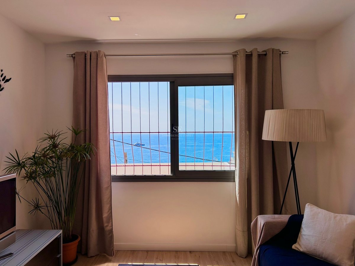 A window with a metal grate framing a blue sea view, flanked by beige curtains, a floor lamp, and a houseplant.