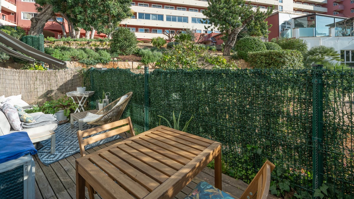 Sun-drenched private terrace featuring a wooden dining set, lounge area with patterned rug, and green privacy fencing.