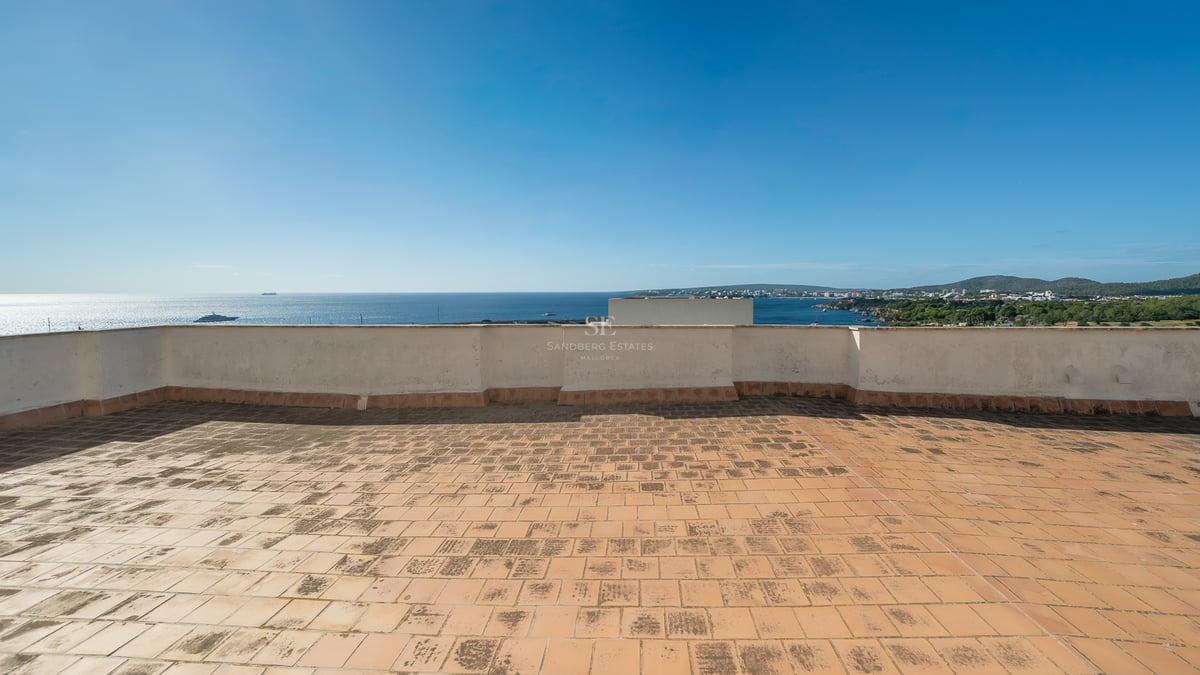 Large rooftop terrace with terracotta tiles overlooking a clear blue sea and coastline under a bright sky.