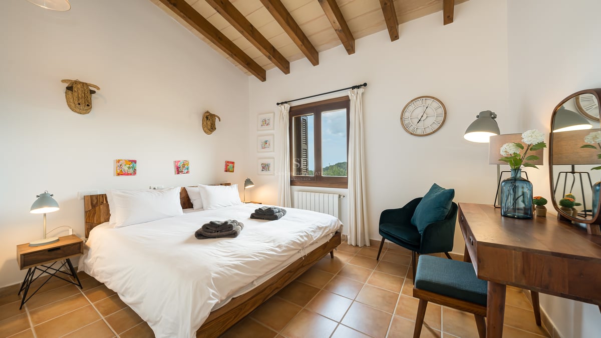Chambre avec sols en terre cuite, poutres en bois apparentes, literie blanche et fauteuil en velours bleu canard.