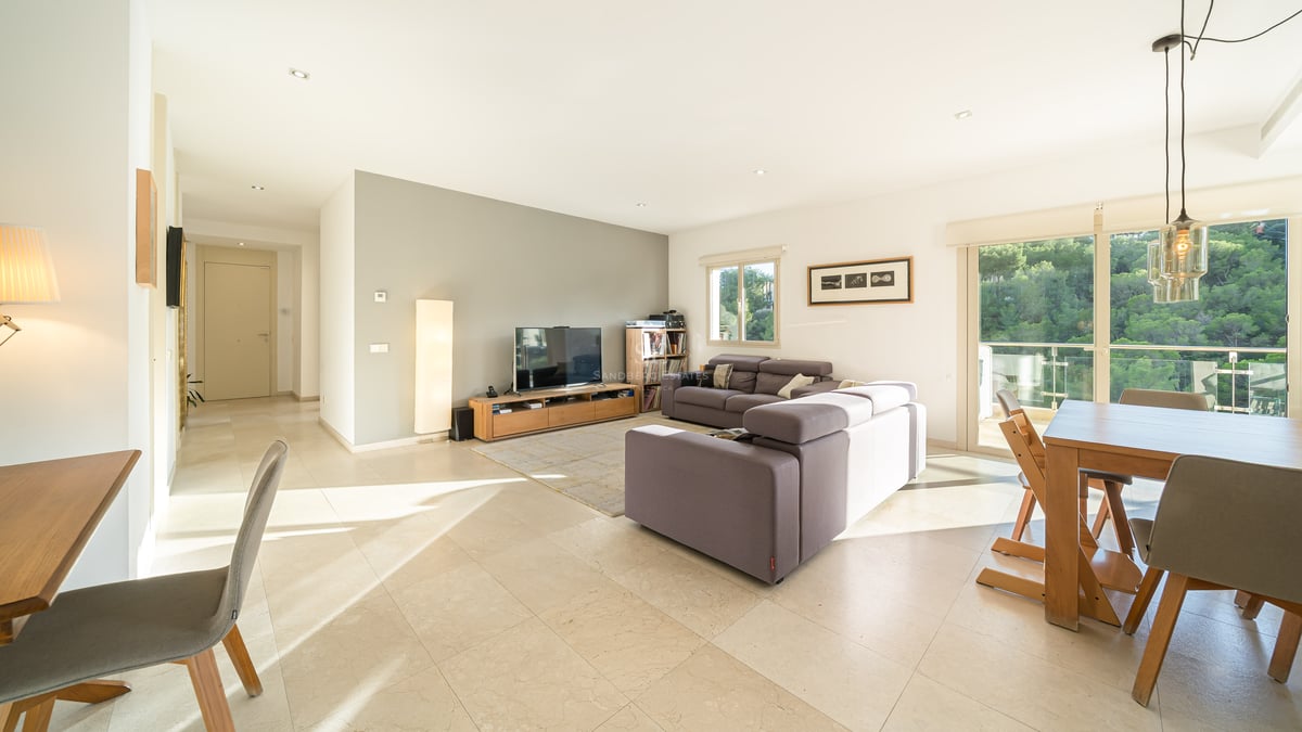 Ljust modernt vardagsrum med grå soffor, matbord i trä och stora fönster som vetter mot en grönskande skog.