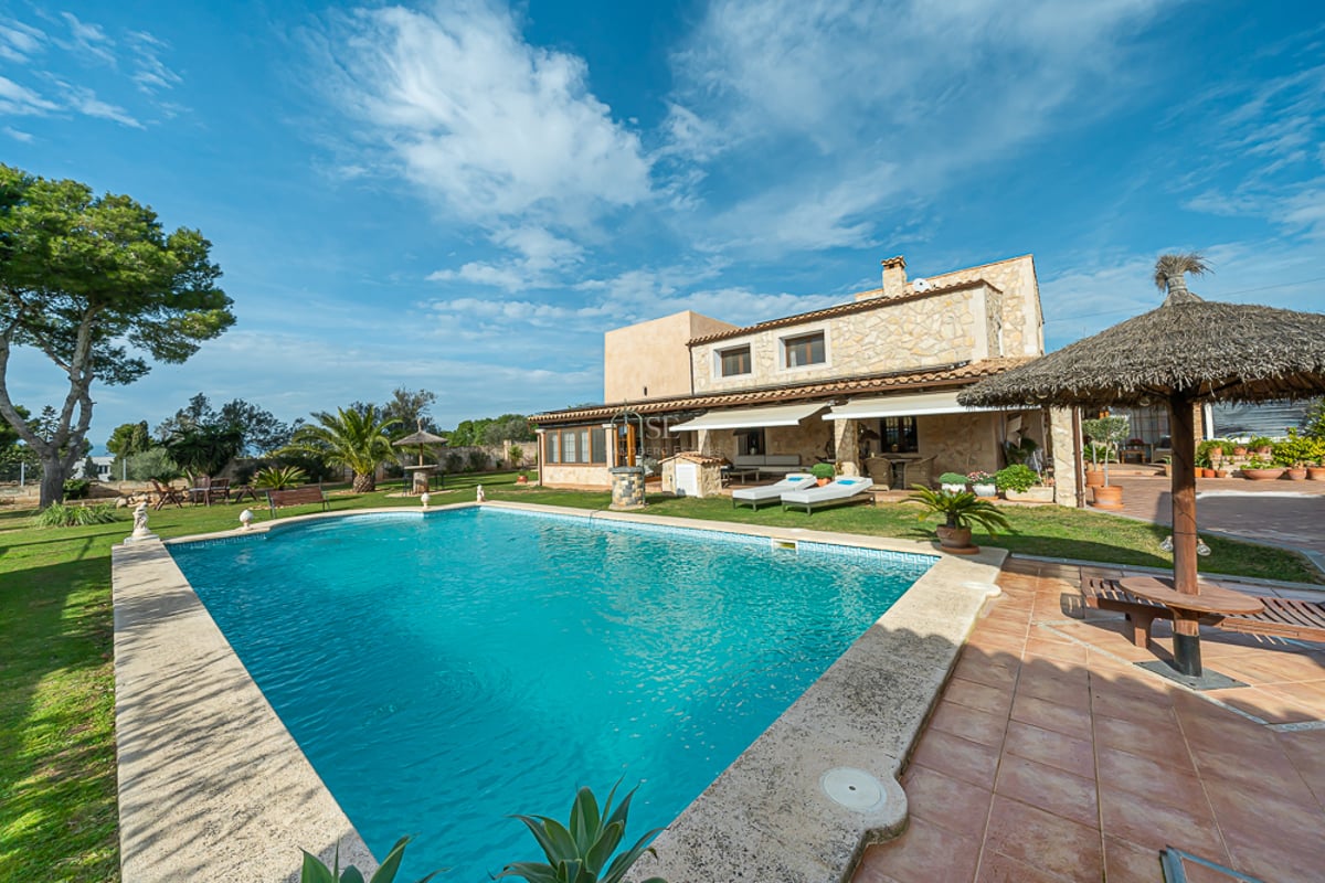 Grande piscine turquoise devant une villa en pierre avec jardin soigné, pelouse et parasols en chaume.