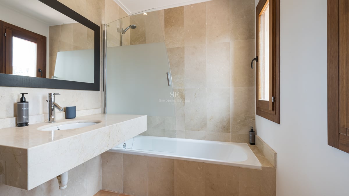 Salle de bain moderne avec carrelage en pierre beige, baignoire blanche, paroi en verre et fenêtre en bois.