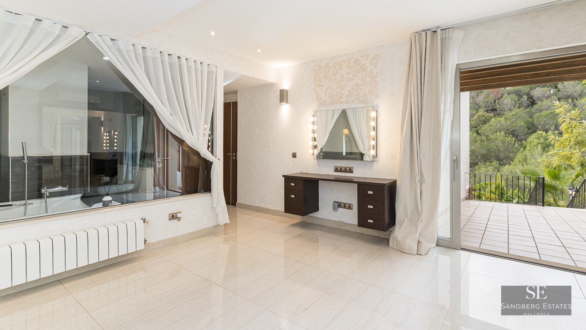 Bright dressing area with a Hollywood mirror vanity, glass wall to a bathroom, and sliding doors to a forest-view terrace.