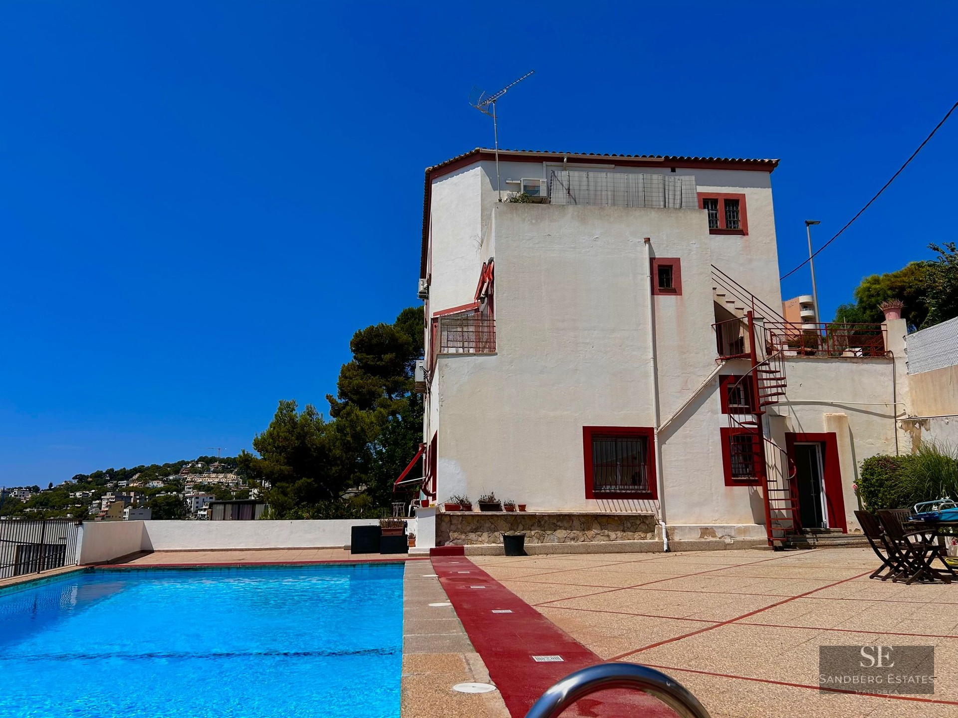 Large outdoor swimming pool in front of a white villa with red trim under a bright blue sky.