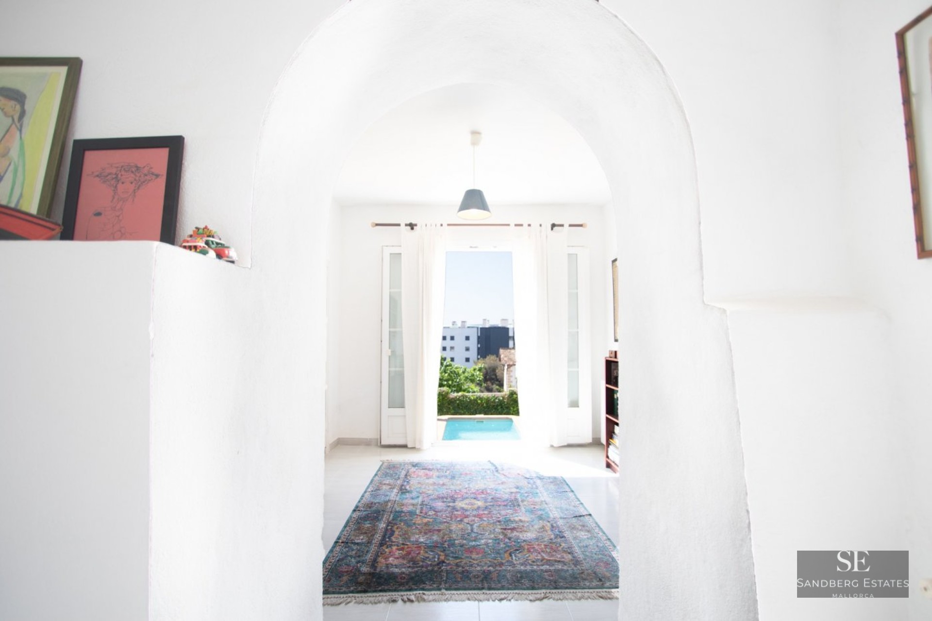 A view through a white plastered archway into a bright room with a colorful oriental rug and a pool visible outside.