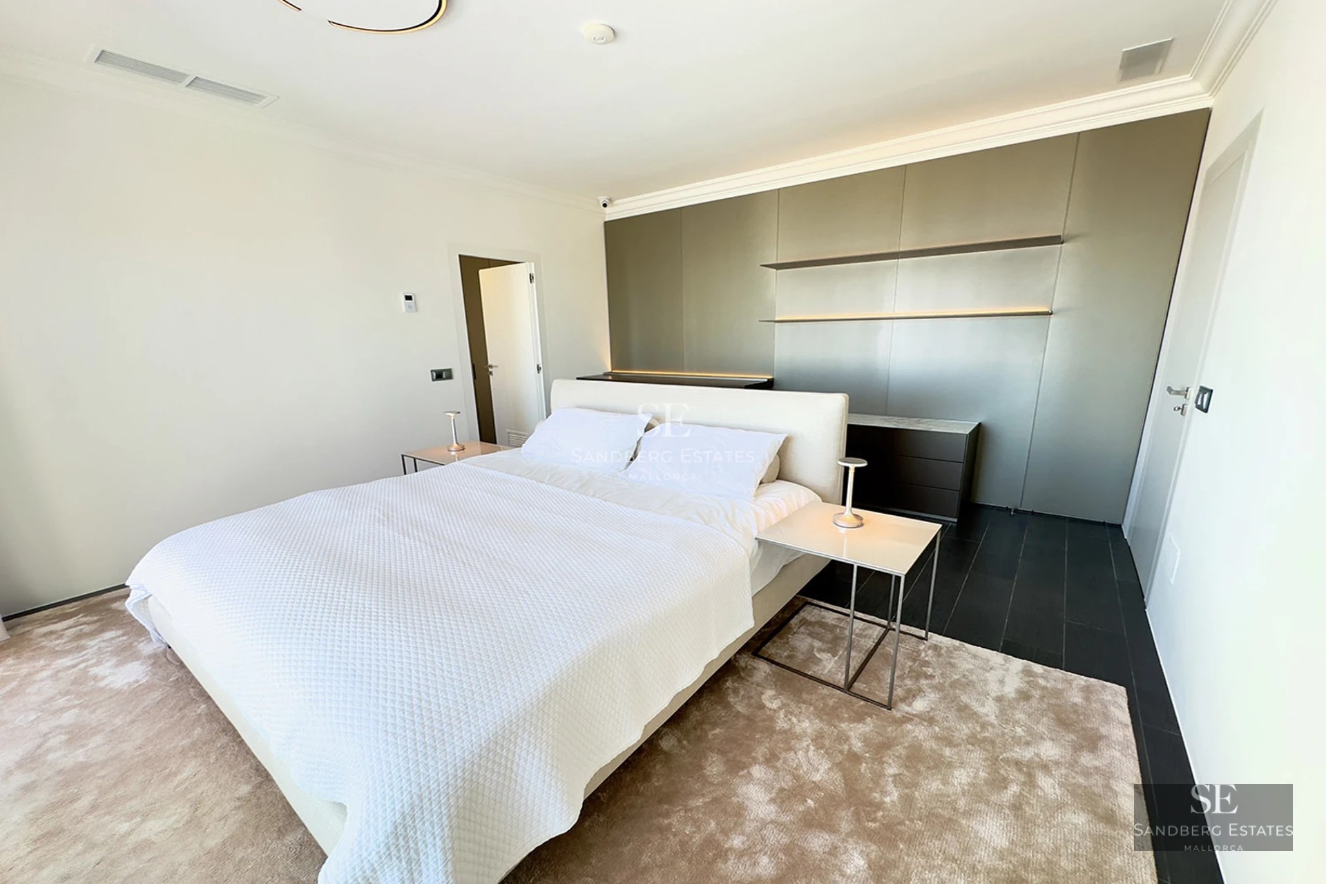 Sleek modern master bedroom featuring a large white bed, plush rug, and a minimalist grey feature wall with lighting.