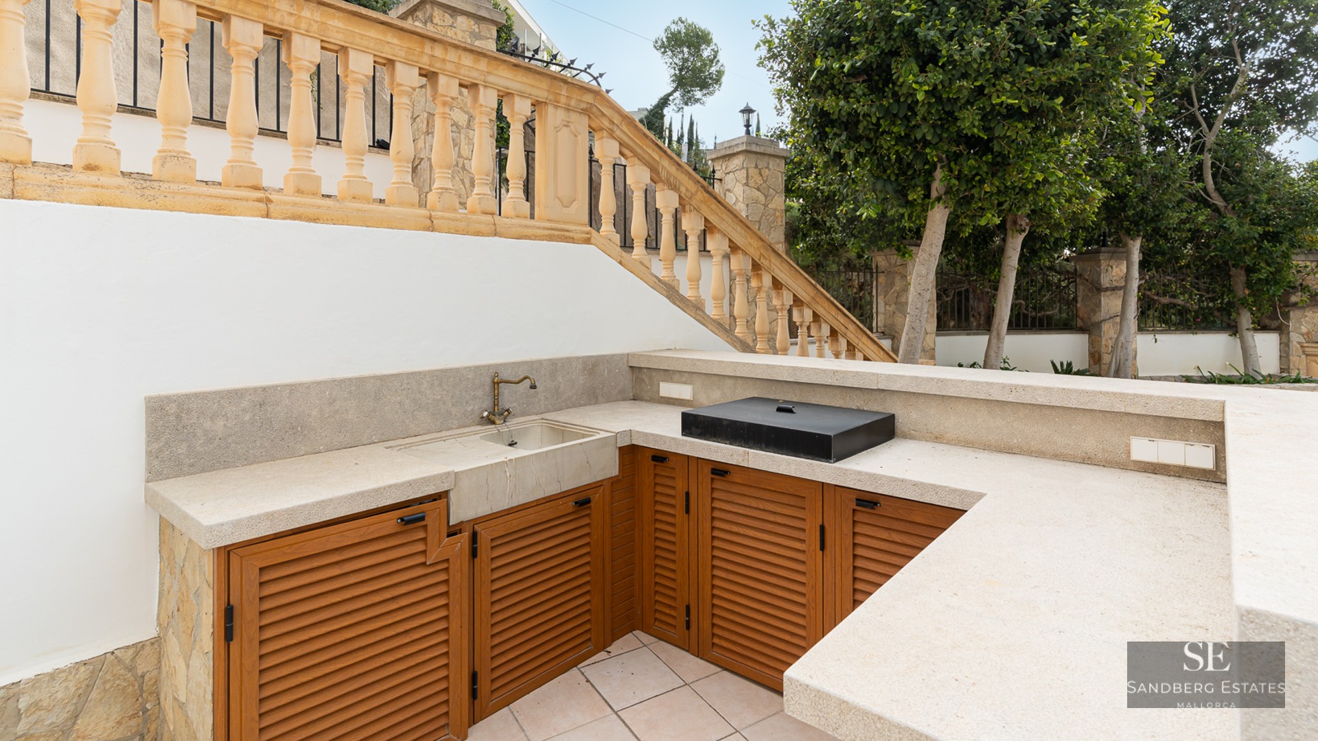Outdoor kitchen with stone countertops, wooden cabinets, and a brass sink next to a classical stone balustrade.