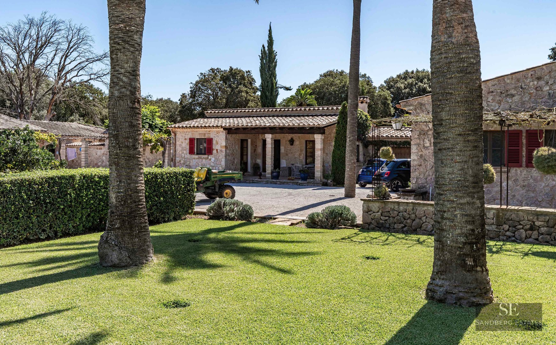 A rustic stone villa with red shutters and a terracotta roof, surrounded by a lush green lawn and palm trees.