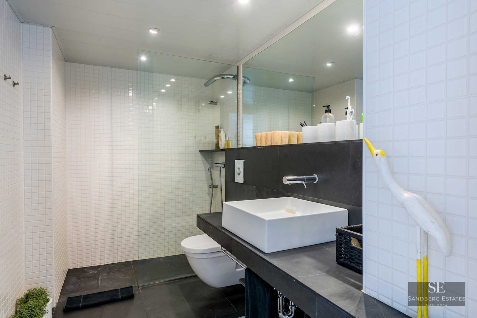 Modern bathroom with white tiled walls, black stone floor, glass shower partition, and a white square vessel sink.