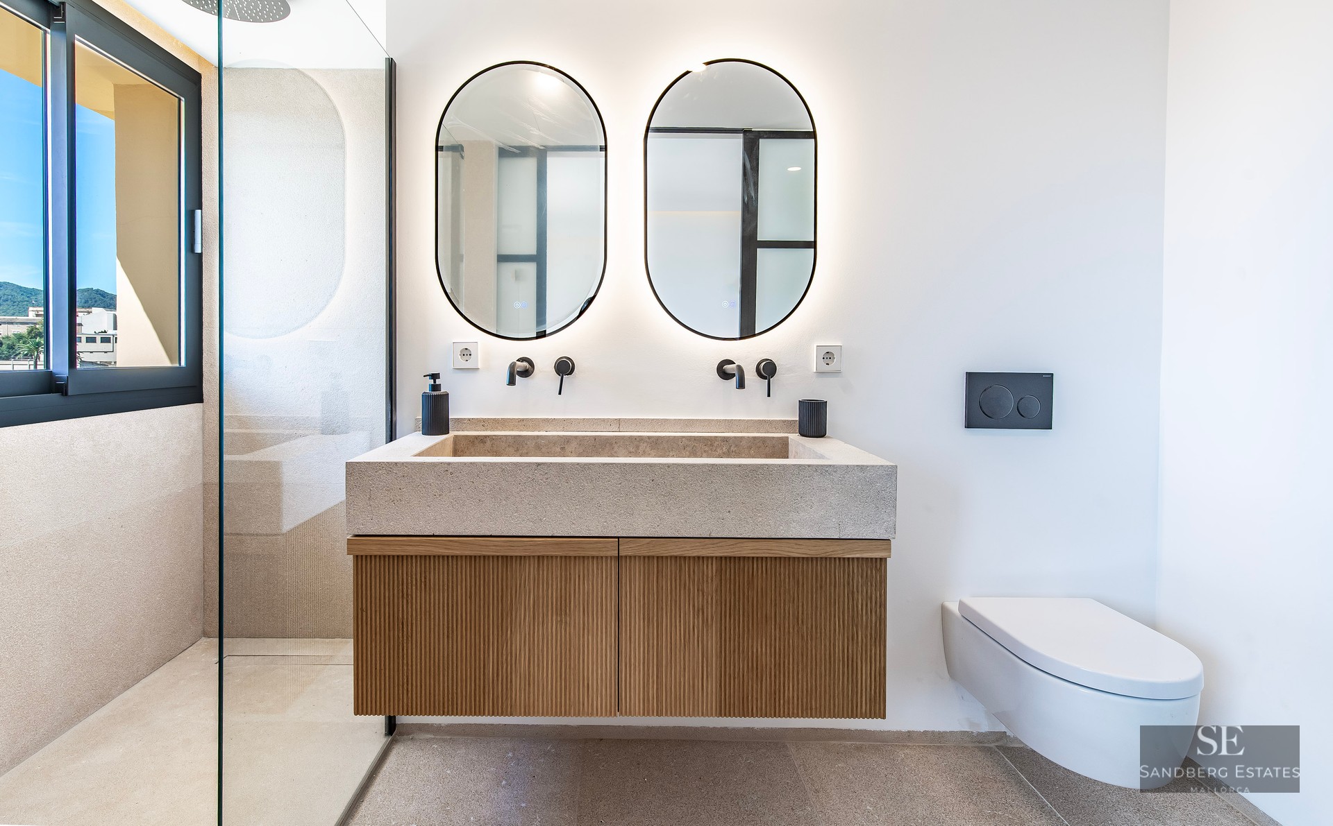 Modern bathroom featuring a stone double vanity, wooden cabinet, two oval backlit mirrors, and a walk-in glass shower.